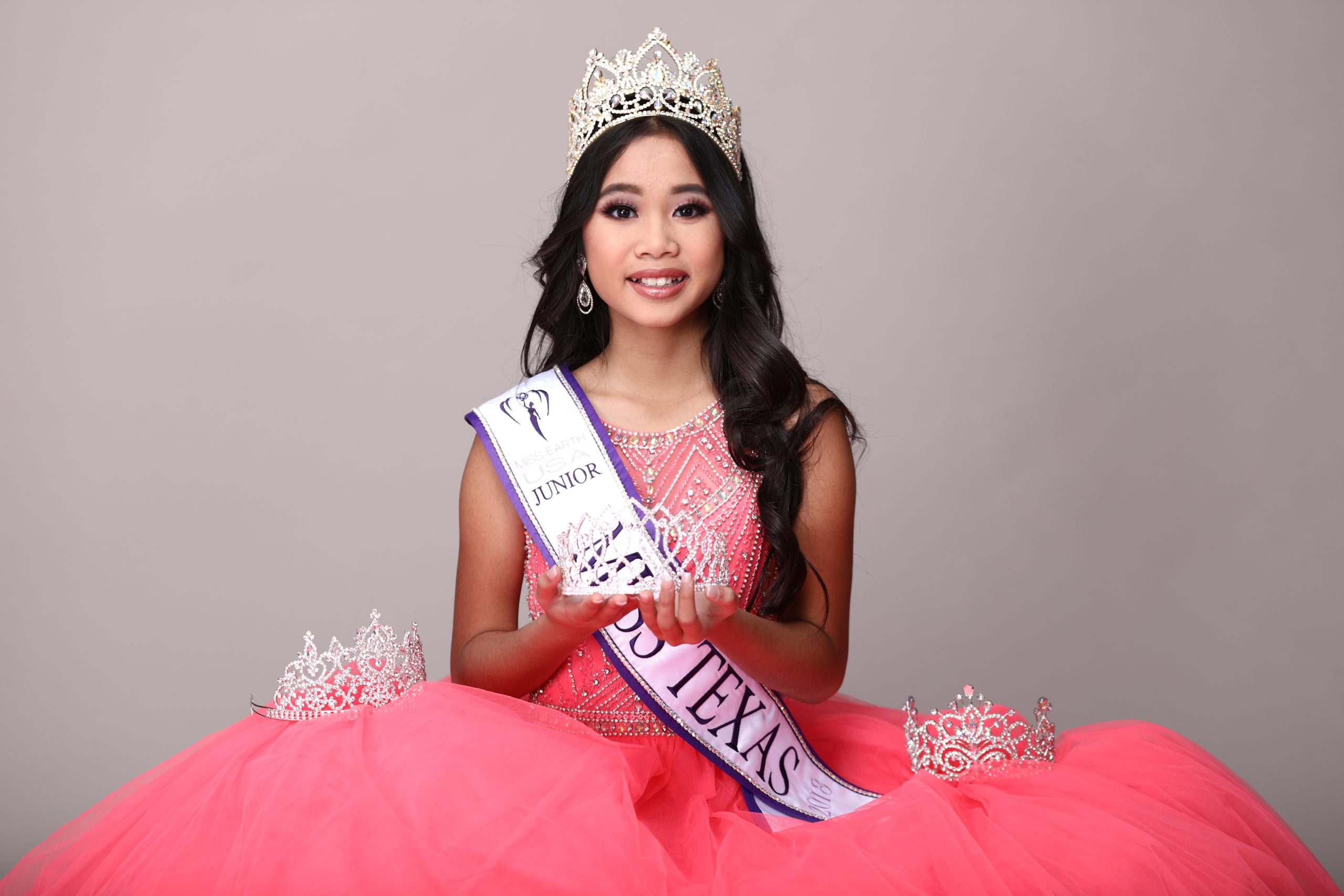 Angelvina, a young woman in a pink dress and crown sits with a sash reading "Junior Miss Texas." She holds a clear crystal award, with additional crowns placed beside her.
