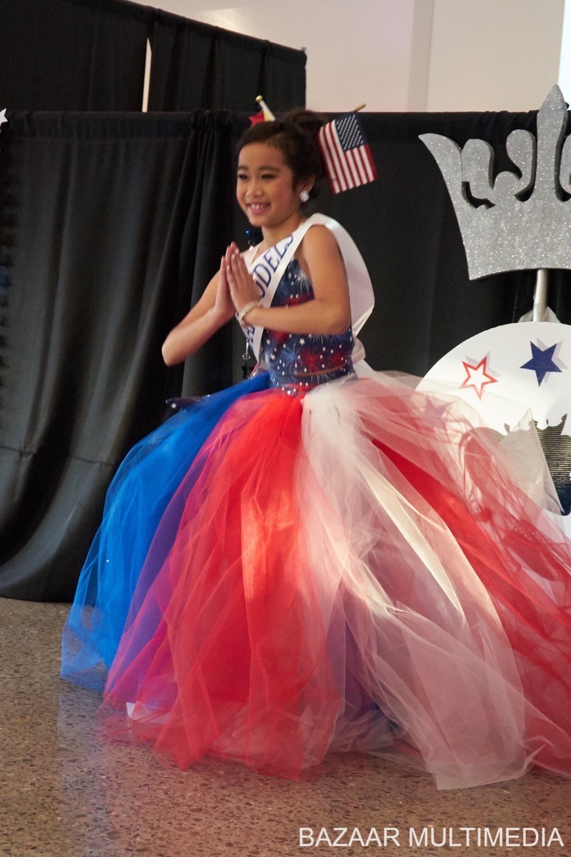 Angelvina Angevlina, A young girl in a red, white, and blue dress with a sash smiles and poses with hands together.