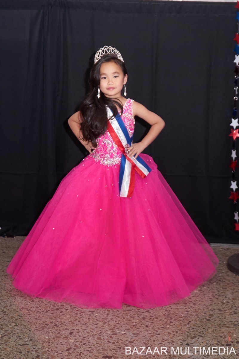 Davina Davina, Young girl in a pink gown and tiara, wearing a sash, standing indoors against a dark backdrop.