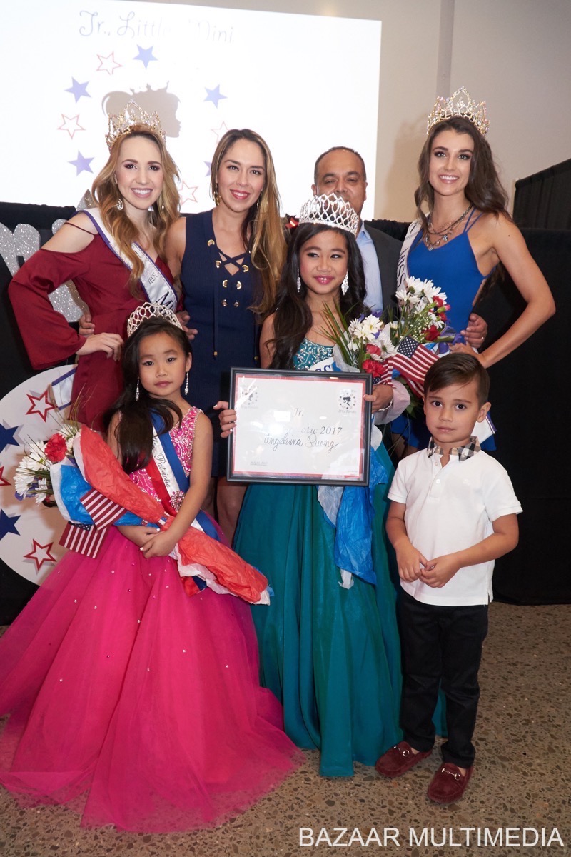 Angelvina Davina A group of people posing in formal attire. Two young girls in gowns hold bouquets. An older child holds a certificate. American flags and a step-and-repeat backdrop are visible.