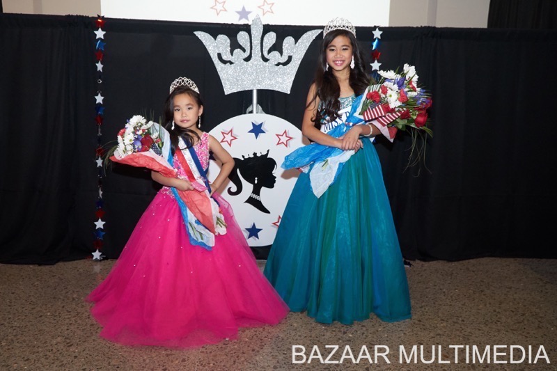 FINAL2374 Two young girls in gowns and tiaras holding flowers stand in front of a decorative backdrop.