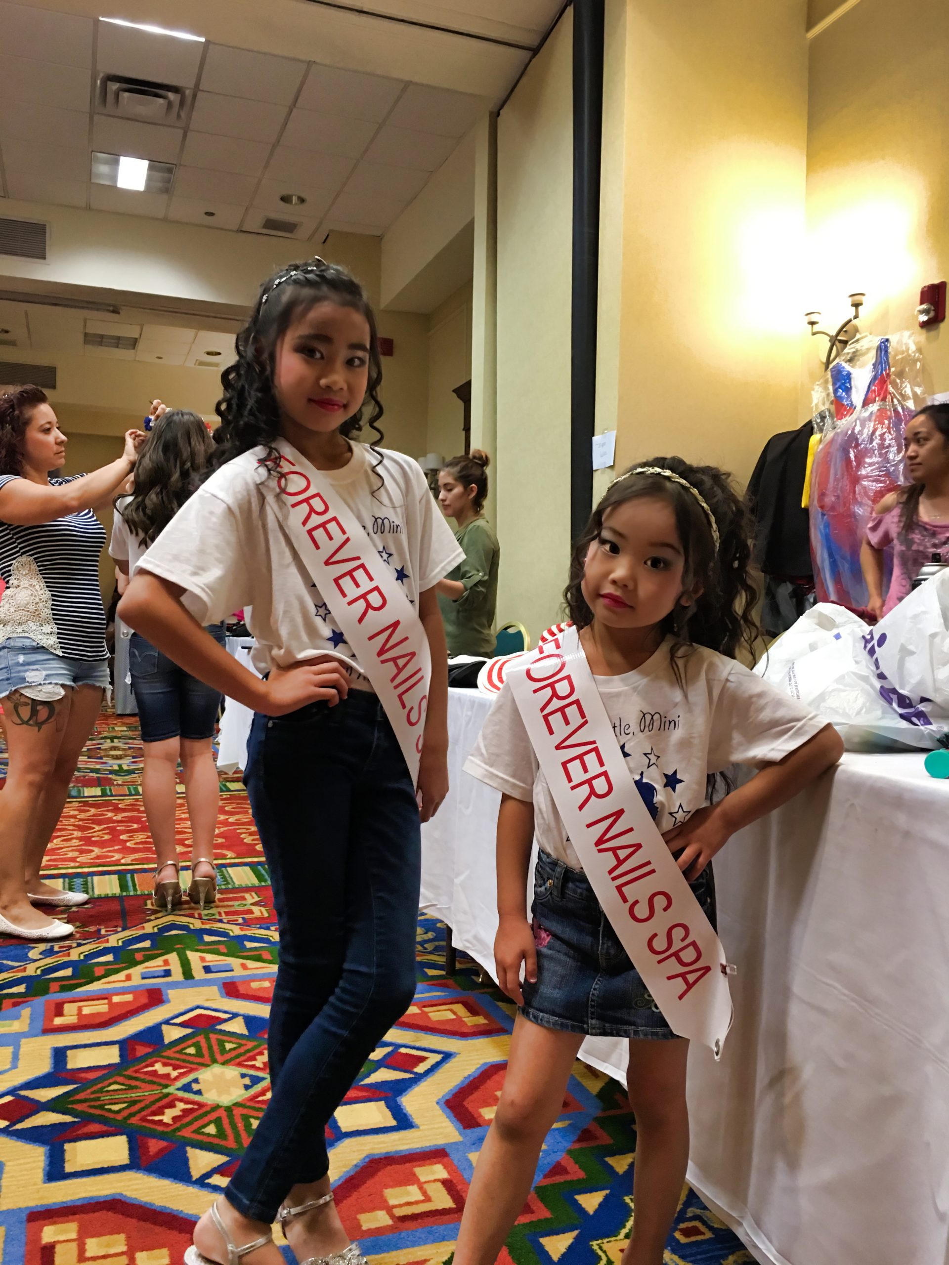 Angelvina Davina; Two young girls wearing "Forever Nails Spa" sashes pose confidently in a room with patterned carpet and people in the background.
