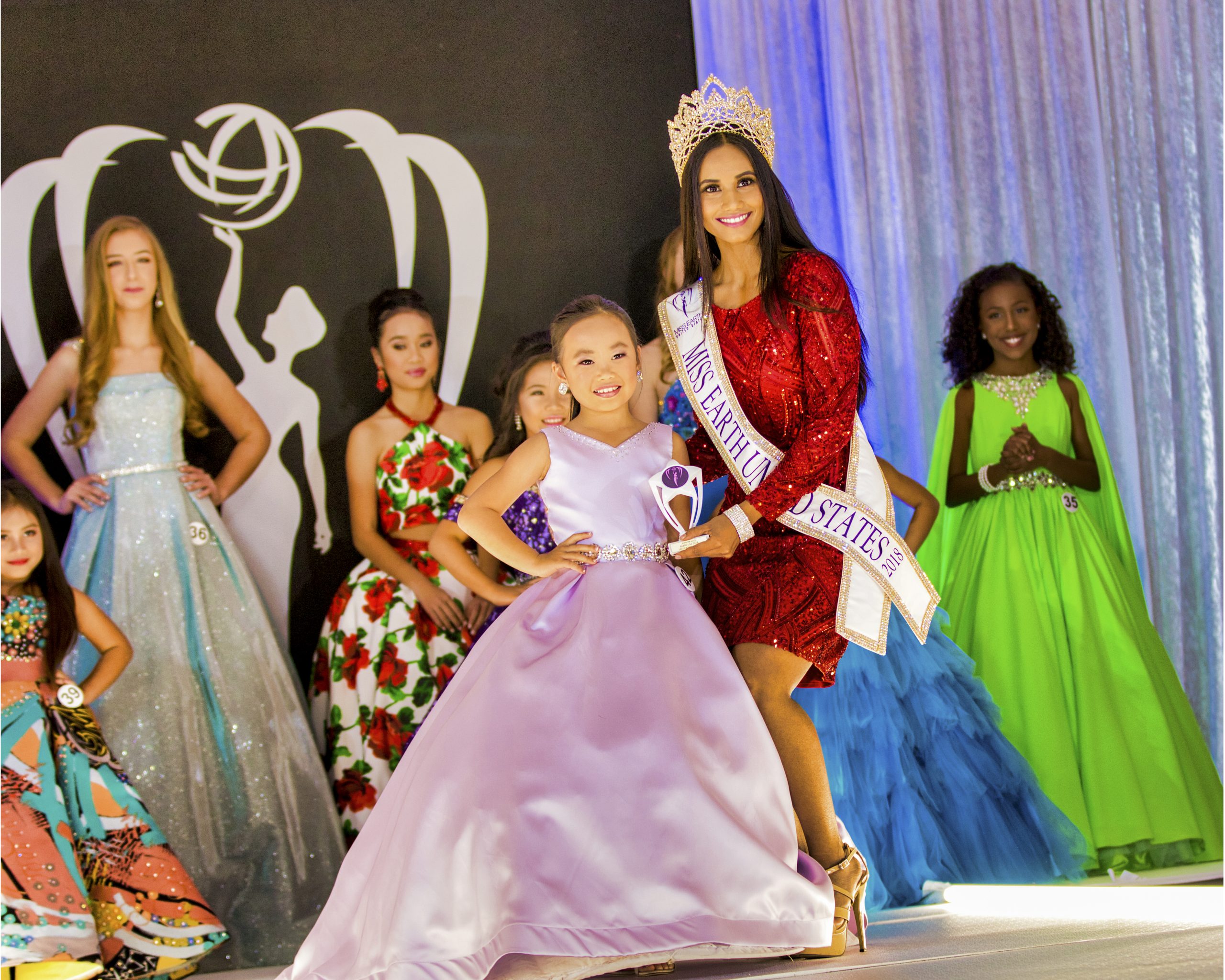 Angelvina Davina Angelvina Davina, A beauty pageant scene with a smiling woman in a red dress and crown beside a young girl in a purple gown holding a trophy. Other contestants in colorful dresses stand in the background.